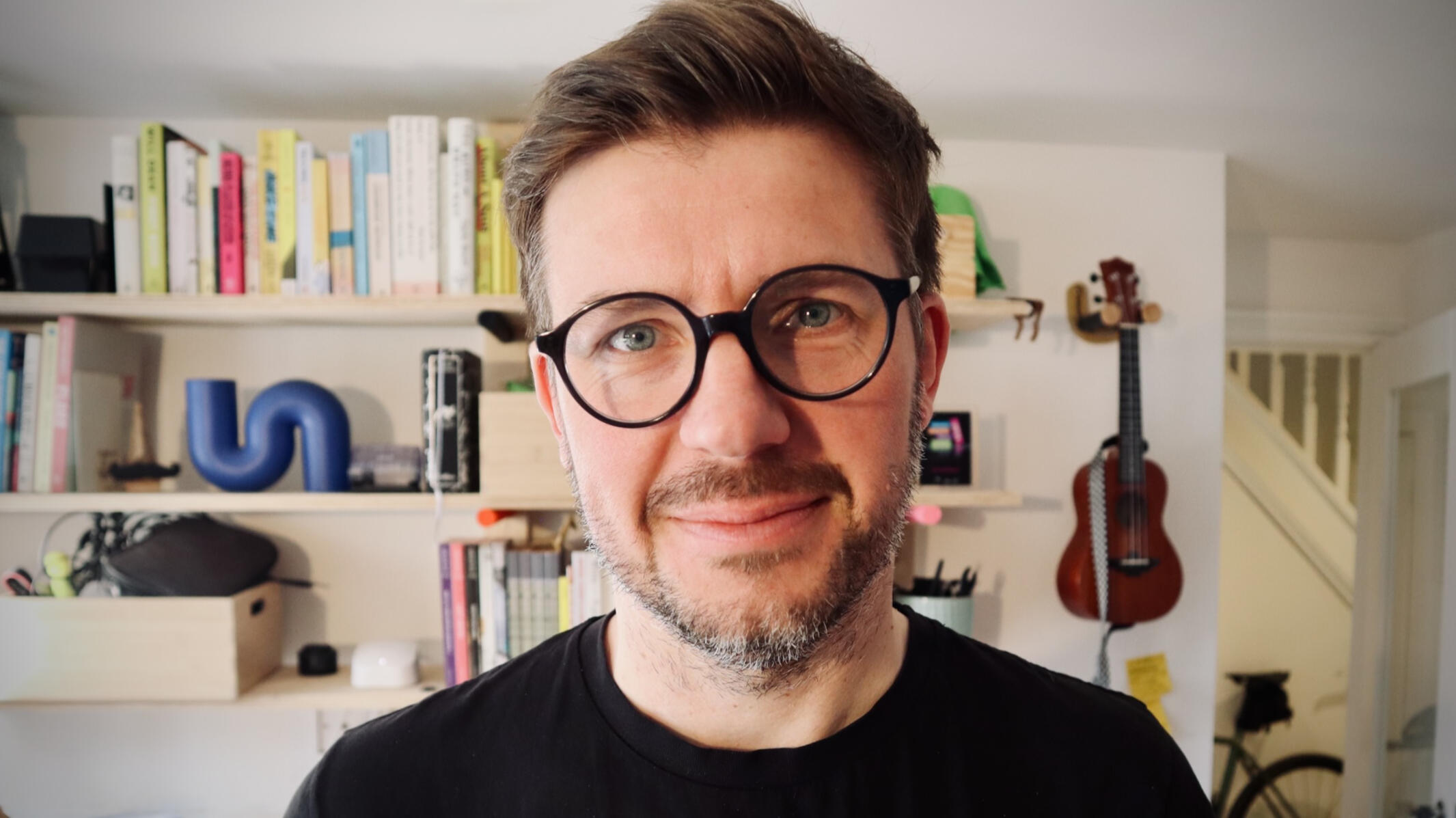 Content designer, Iain Broome from Very Meta Iain Broome headshot with an office background behind him. He is wearing a black t-shirt, glasses and a smile that suggests he may be tired of trying to take a good profile picture. In the background, slightly out of focus, there are shelves with books, or